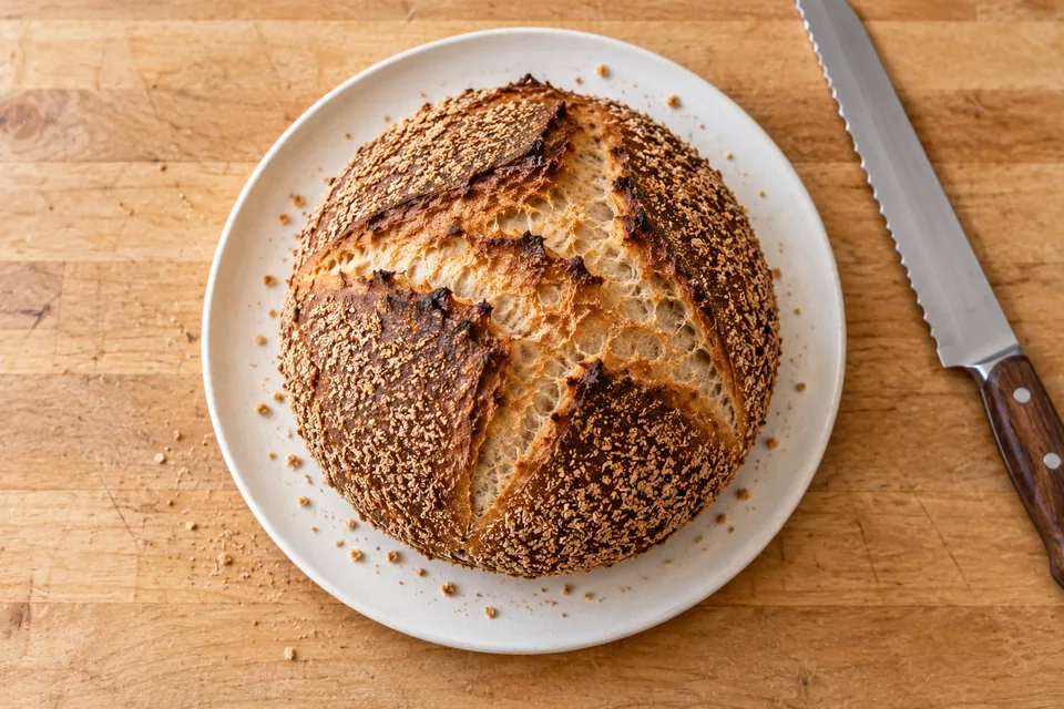 A whole bran-coated round loaf on a plain white plate, photographed from above on a wooden counter.