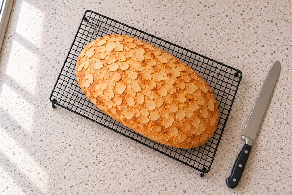 One oval loaf covered with golden almond flakes rests diagonally on a black cooling rack, viewed from above.
