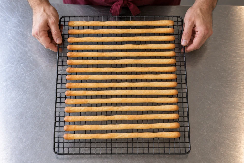 Fifteen long golden plain breadsticks from one tray cool in parallel rows on a wire rack.