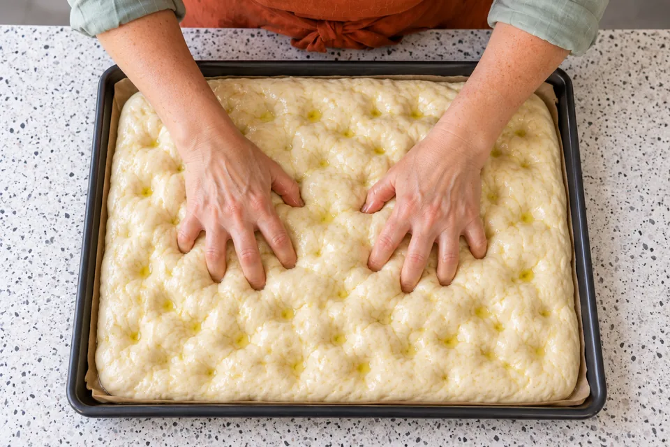 Fingertips dimple plain focaccia dough with a light oil and brine coating.