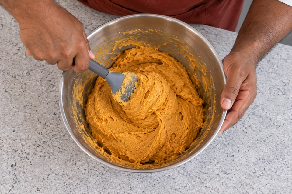 Grey spatula folds thick orange cookie dough with no flour streaks.